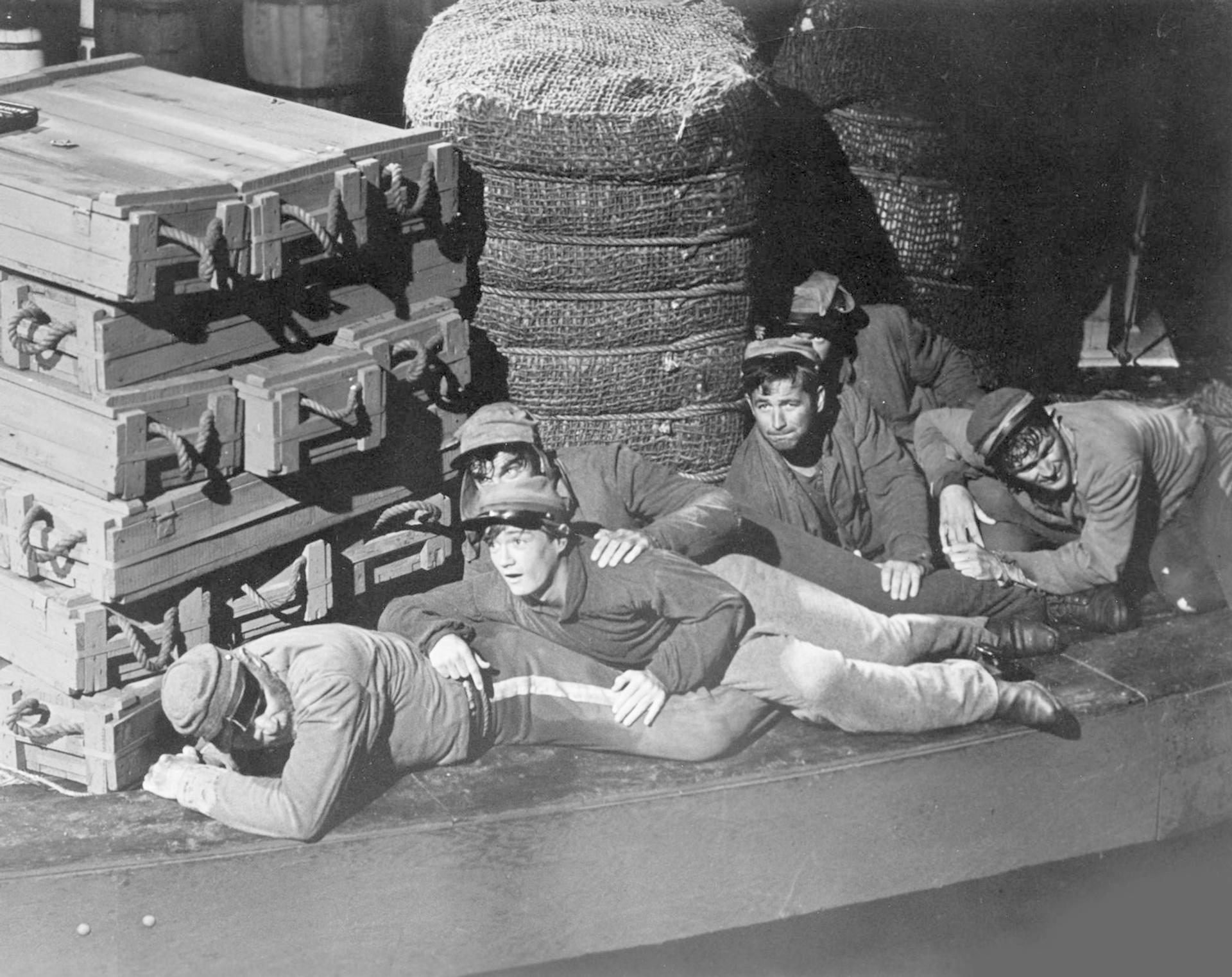 Schwarz-Weiß-Foto von Männern in Uniform, die auf einem Boot liegen. Auf der linken Seite sind Holzkisten gestapelt. Dahinter befindet sich eine große Webstruktur.