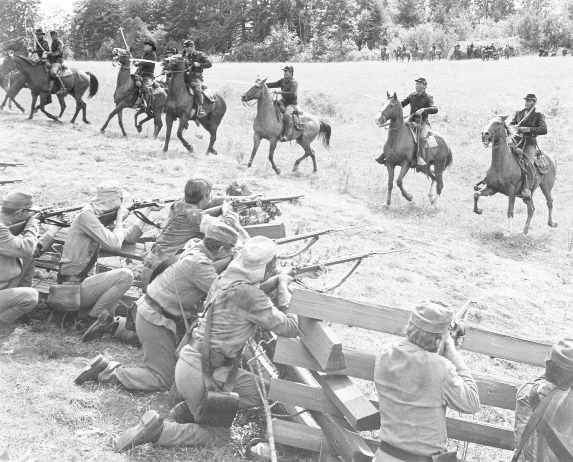 Nachstellung des Bürgerkriegs: Kavallerie greift Infanterie hinter einem Holzzaun an. Soldaten feuern Gewehre ab; Pferde galoppieren.