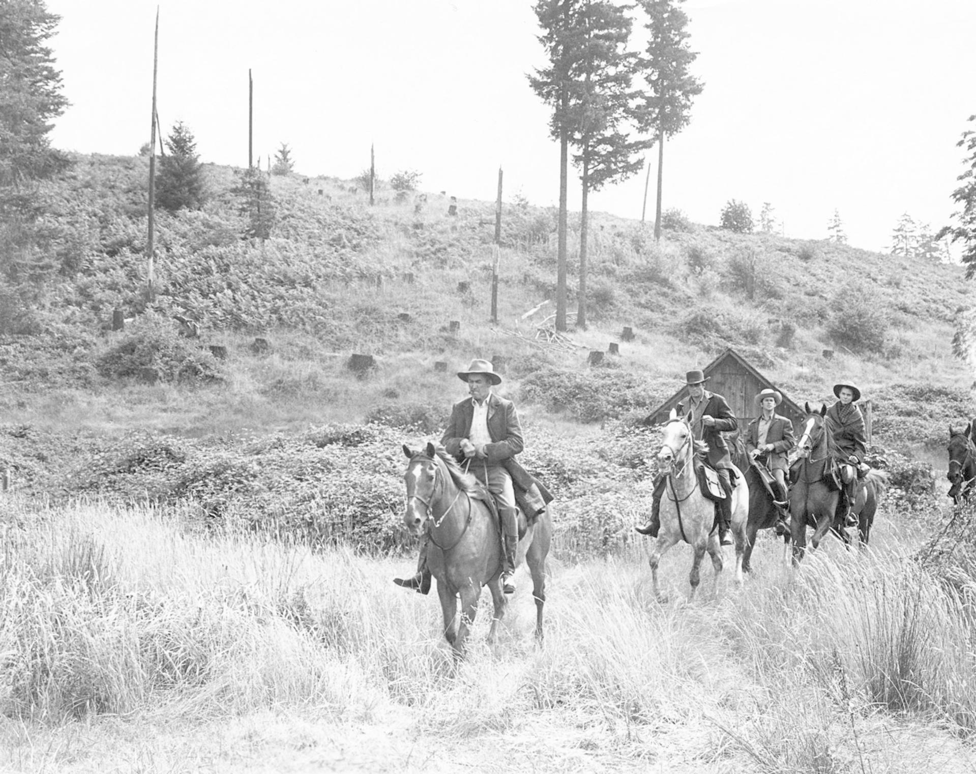 Fünf Personen reiten auf Pferden durch ein Feld. Im Hintergrund befinden sich eine kleine Hütte und Bäume. Die Szene ist in Graustufen gehalten.