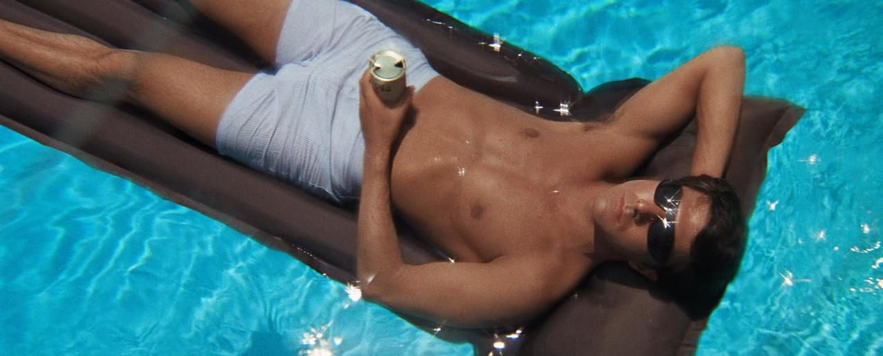 Ein Mann mit Sonnenbrille schwimmt auf einer Poolliege und hält eine Dose. Er trägt Badehosen. Klares blaues Wasser umgibt ihn.