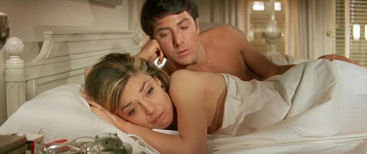 Ein Mann und eine Frau liegen im Bett. Die Frau schaut in die Kamera; der Mann schaut weg. Weiße Bettwäsche und ein Kopfteil sind sichtbar.