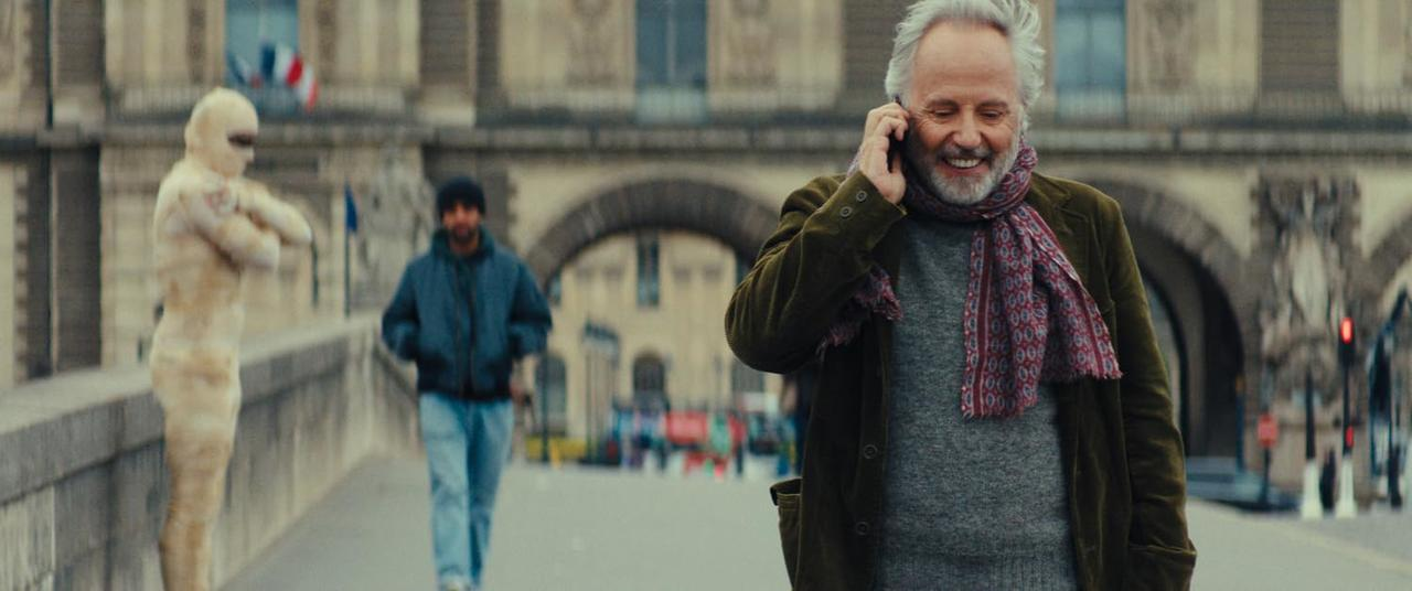 Mann am Telefon, lächelnd. Ein anderer Mann geht dahinter. Eine in Stoff gehüllte Person steht links. Im Hintergrund ist ein Torbogen.