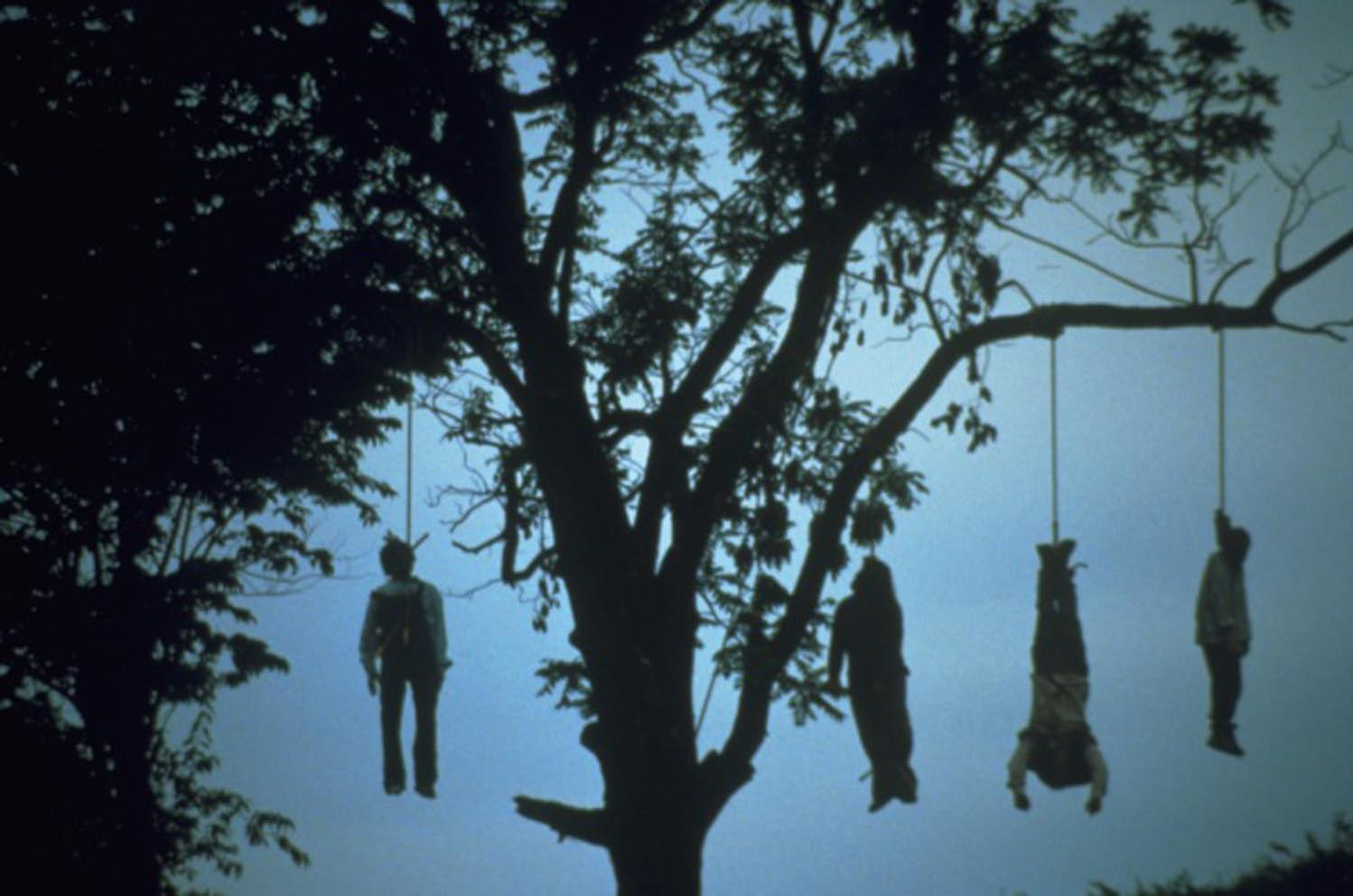Silhouette eines großen Baumes mit mehreren Figuren, die an Seilen hängen, vor blauem Himmel. Dunkle, ominöse Szene.