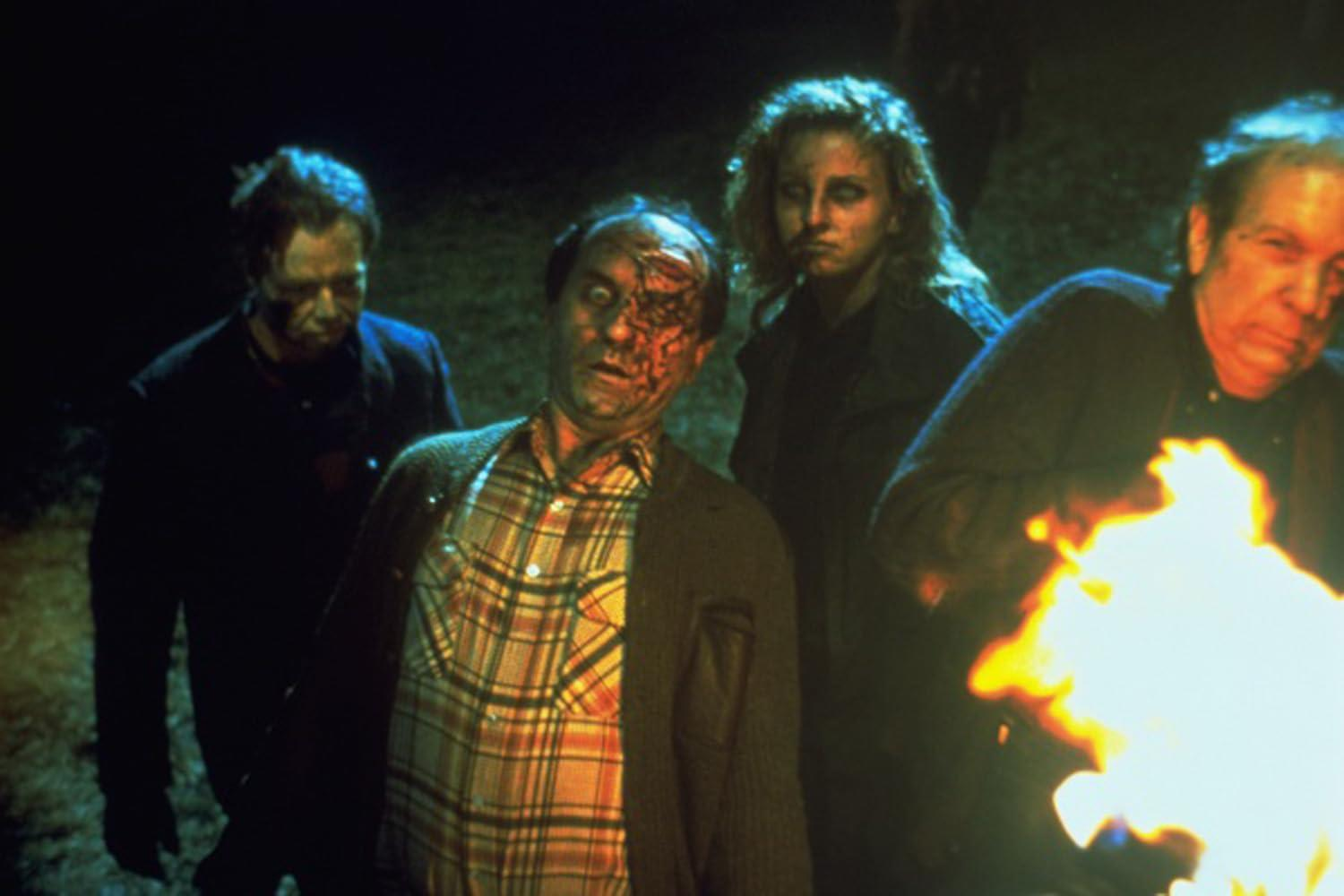 Vier Personen in einer dunklen Szene, einige mit zombieartigen Merkmalen, in der Nähe eines Feuers. Einer hat ein verbranntes Gesicht, ein anderer ein kariertes Hemd.