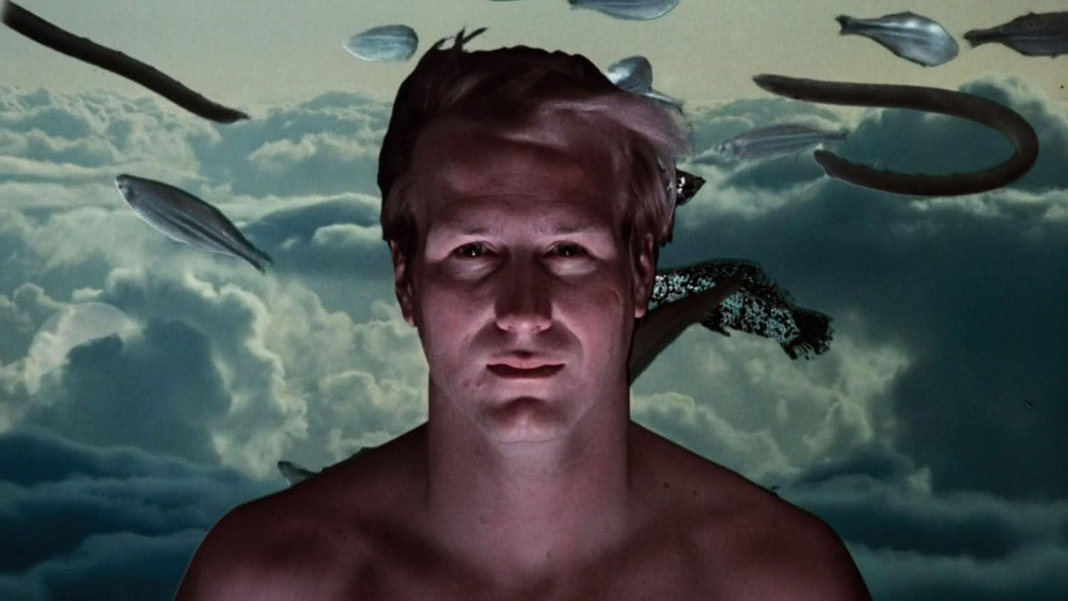 Ein Mann mit Licht im Gesicht, Fische und andere Objekte schweben am Himmel. Im Hintergrund sind Wolken.