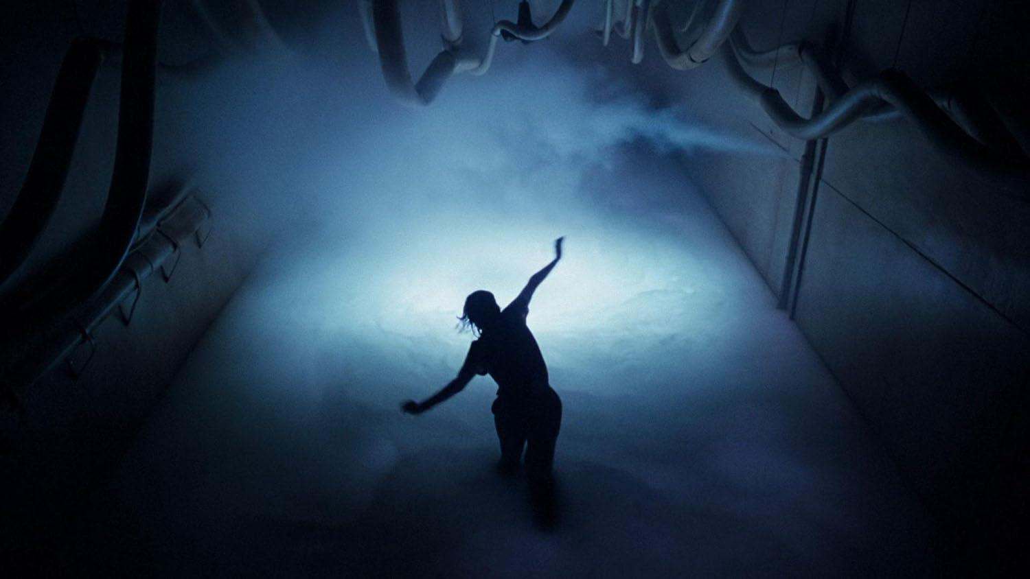 Silhouette einer Person mit erhobenen Armen in einem nebligen, industriellen Raum. Überkopfrohre und Wände sind sichtbar. Blautöne dominieren.