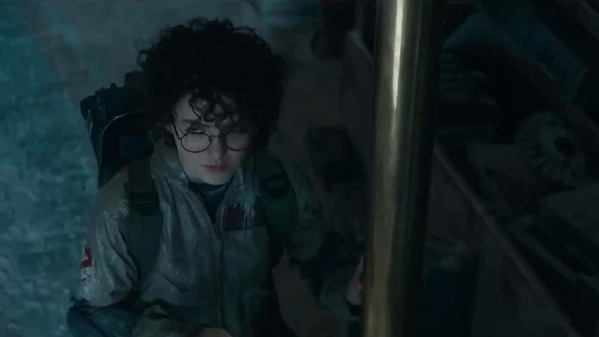 Eine Person in einem Ghostbusters-Anzug mit lockigem Haar und Brille blickt nach unten. Der Hintergrund zeigt einen dunklen Raum mit einer Metallstange.