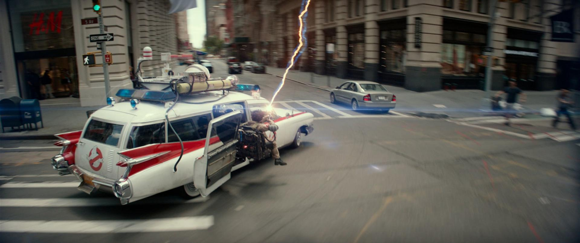 Das Ecto-1-Auto von Ghostbusters ist in Aktion, ein Strahl schießt daraus auf einer Straße in der Stadt. Gebäude und andere Autos sind sichtbar.