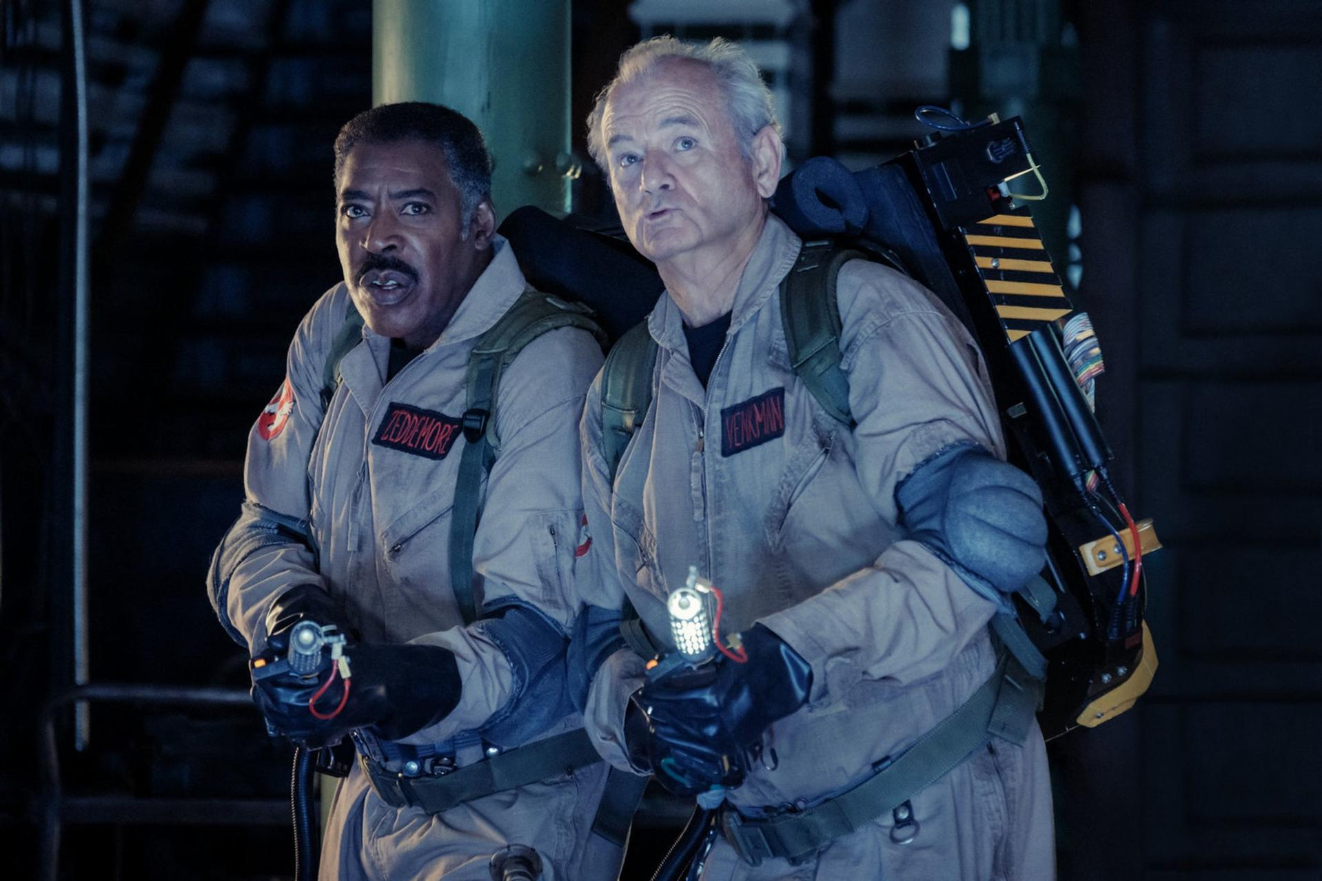 Zwei Männer in Ghostbusters-Uniformen halten Protonenpacks. Einer ist schwarz, der andere ist weiß. Beide sehen besorgt aus. Dunkler Hintergrund.