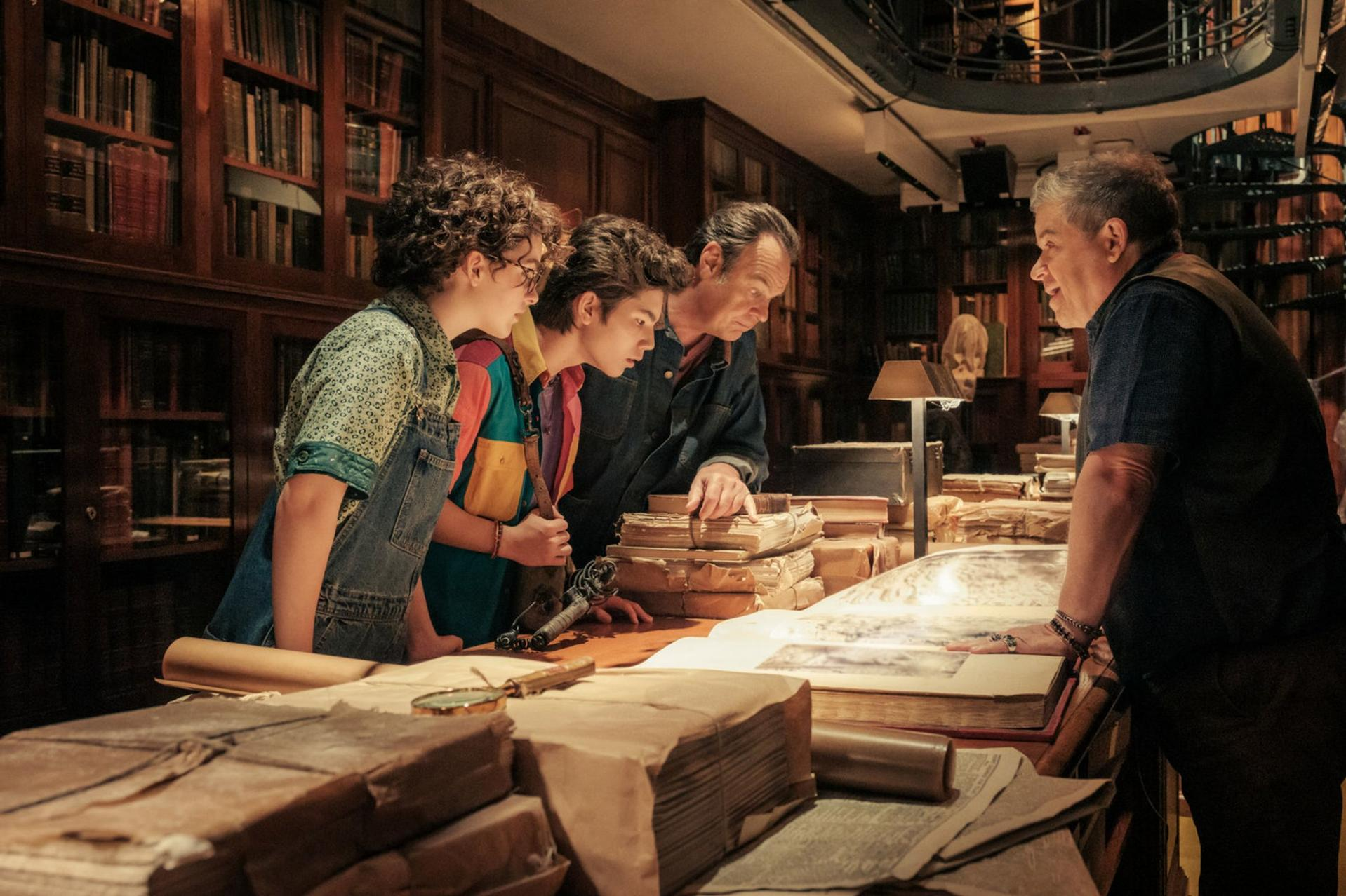Eine Gruppe von Leuten betrachtet Bücher in einer Bibliothek. Bücherregale im Hintergrund. Ein Mann spricht. Auf dem Tisch liegen weitere Bücher und Gegenstände.