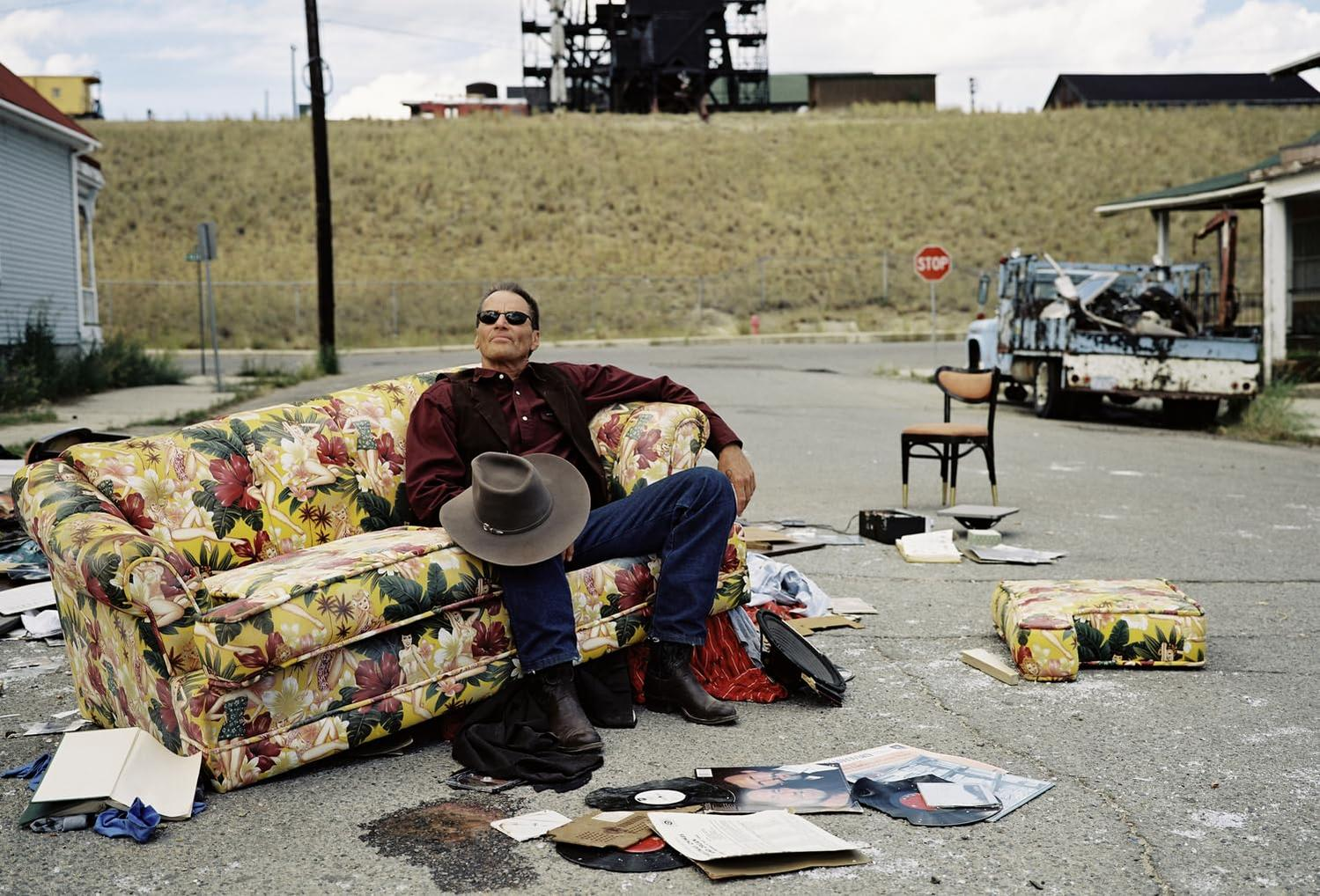 Mann mit Sonnenbrille sitzt auf einem Blumensofa auf der Straße. Ein Hut liegt auf seinem Schoß. Schallplatten, ein Stuhl und Trümmer sind verstreut.