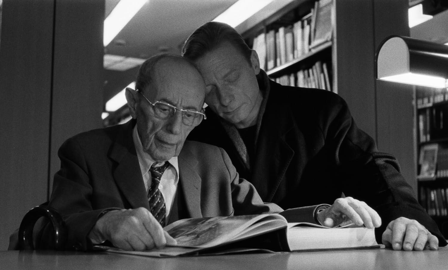 Zwei Männer lesen zusammen in einer Bibliothek ein Buch. Einer lehnt seinen Kopf an die Schulter des anderen.