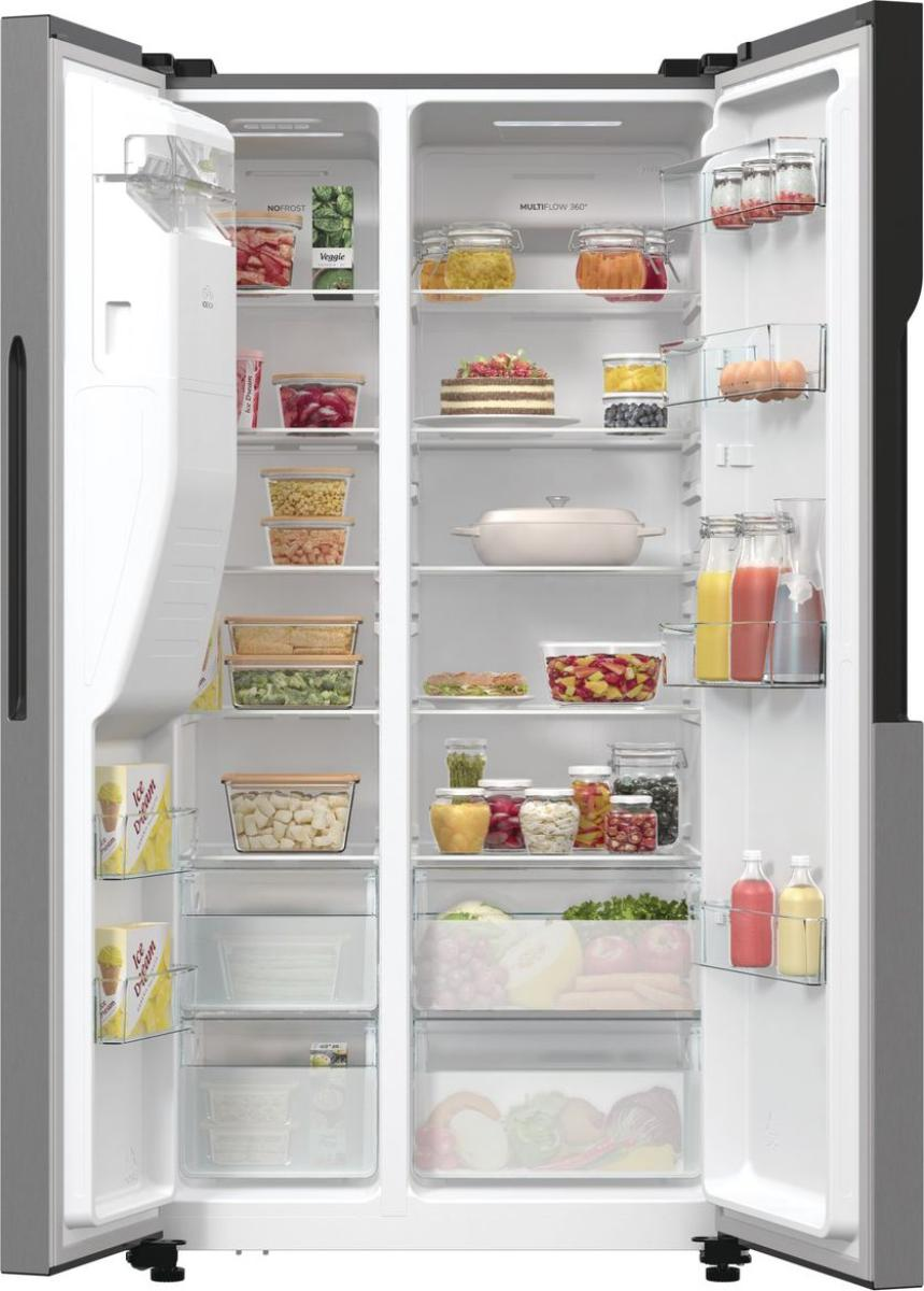 Otwarta lodówka wypełniona jedzeniem. Widoczne słoiki, pojemniki, ciasto, warzywa, owoce i soki.