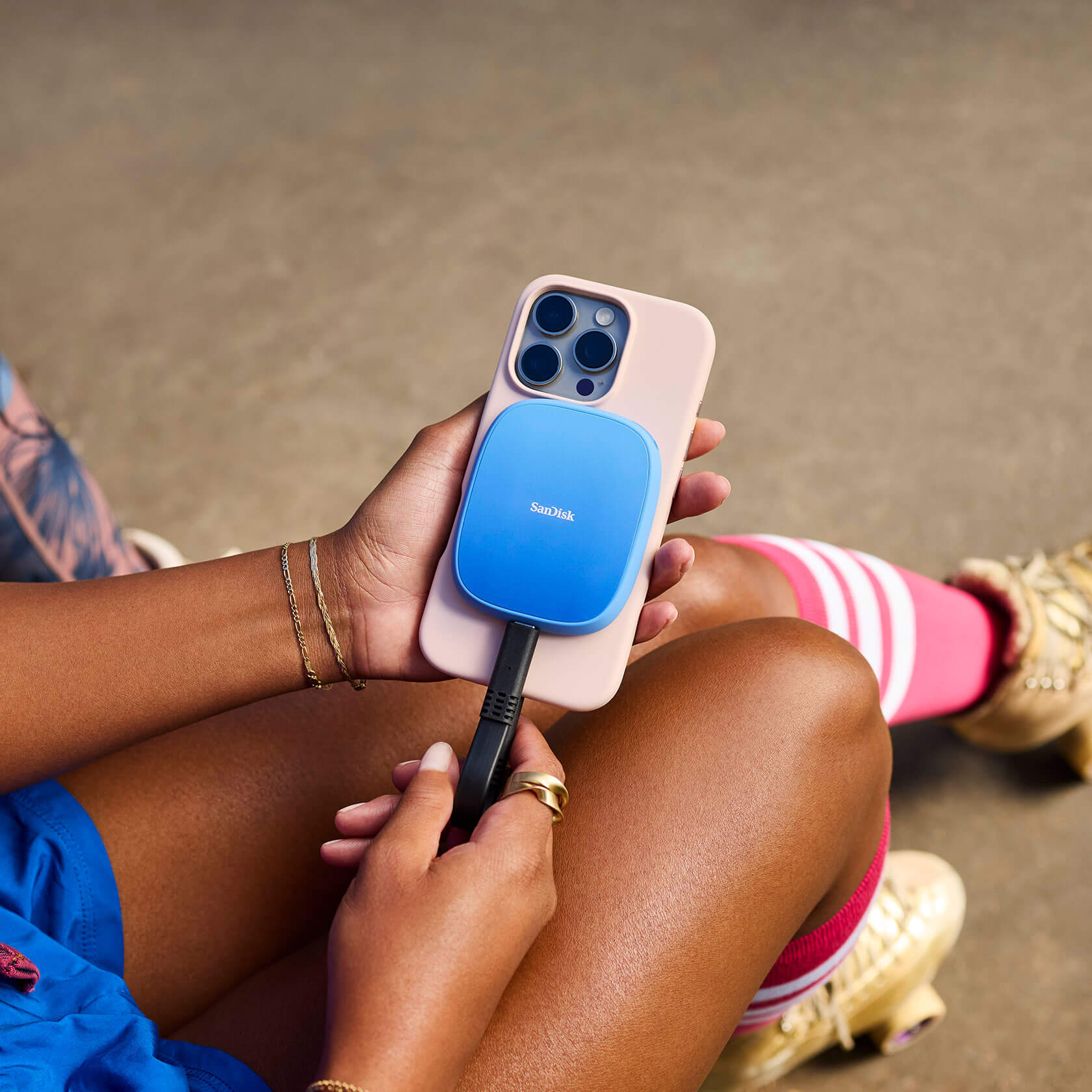 Persona che tiene in mano un telefono con un dispositivo SanDisk collegato. Custodia rosa, dispositivo blu, calzini rosa, gioielli d'oro.