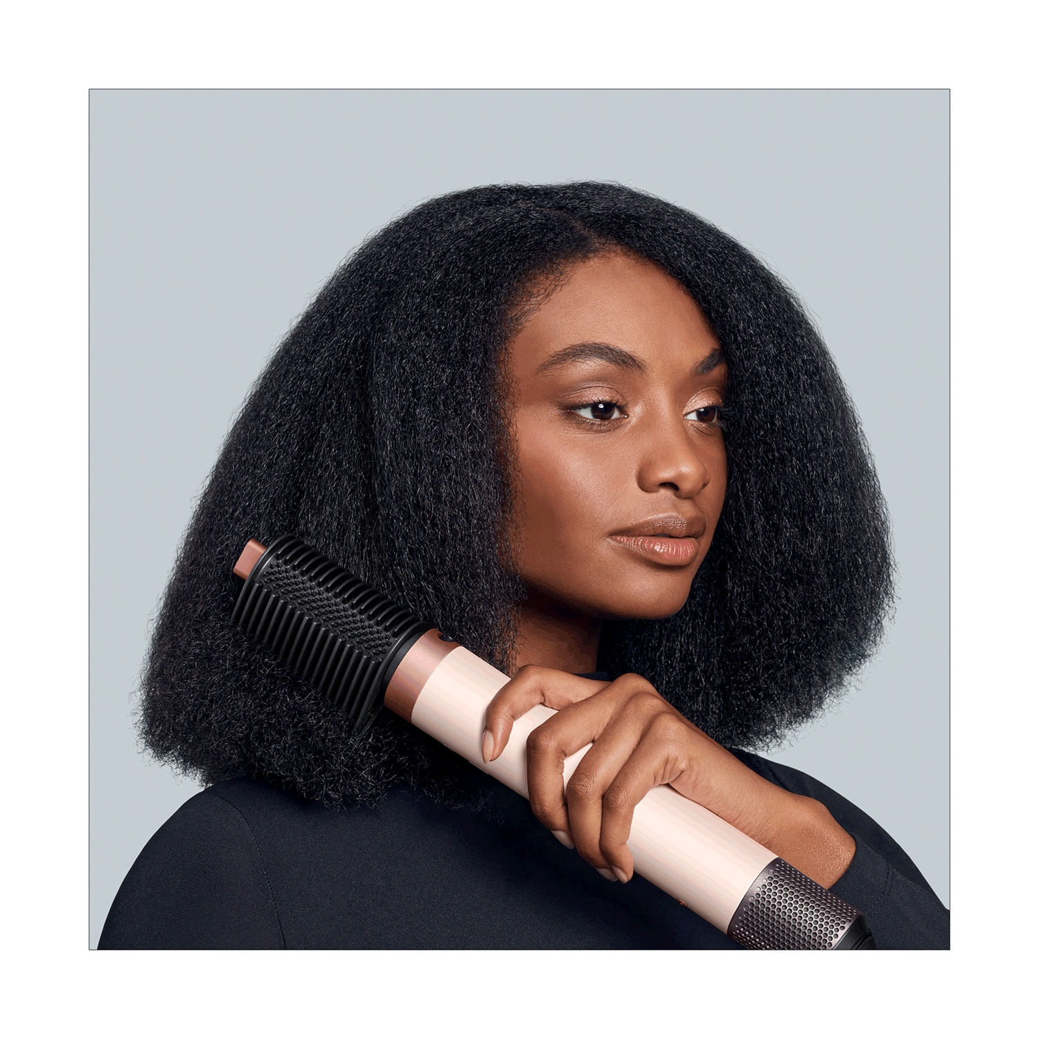 Una donna con i capelli scuri tiene in mano uno strumento per lo styling dei capelli. Lo strumento è bianco e oro rosa, con una testina nera. Indossa un top nero.