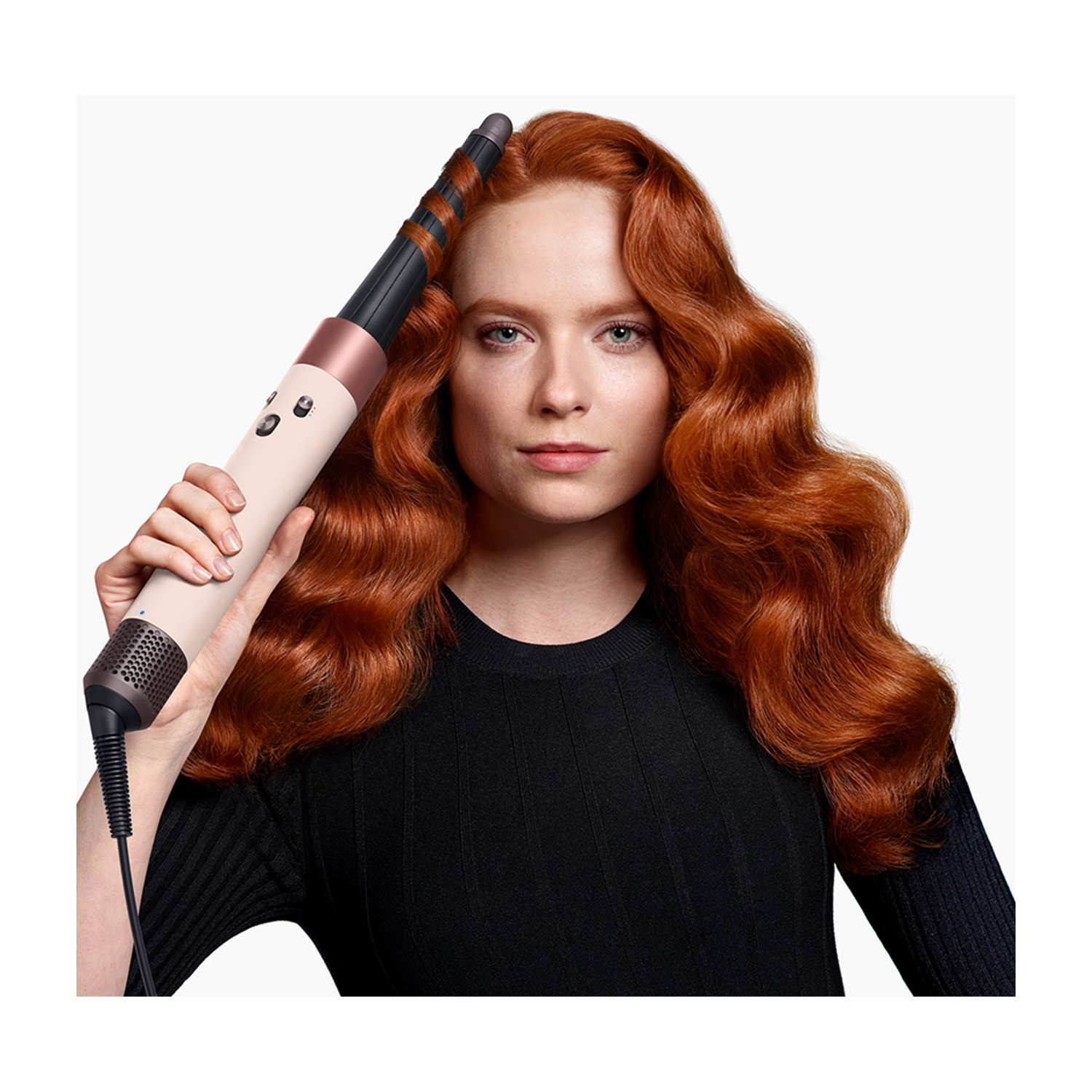 Una donna con capelli rossi e mossi tiene in mano un arricciacapelli. Indossa una camicia nera. Lo sfondo è bianco.
