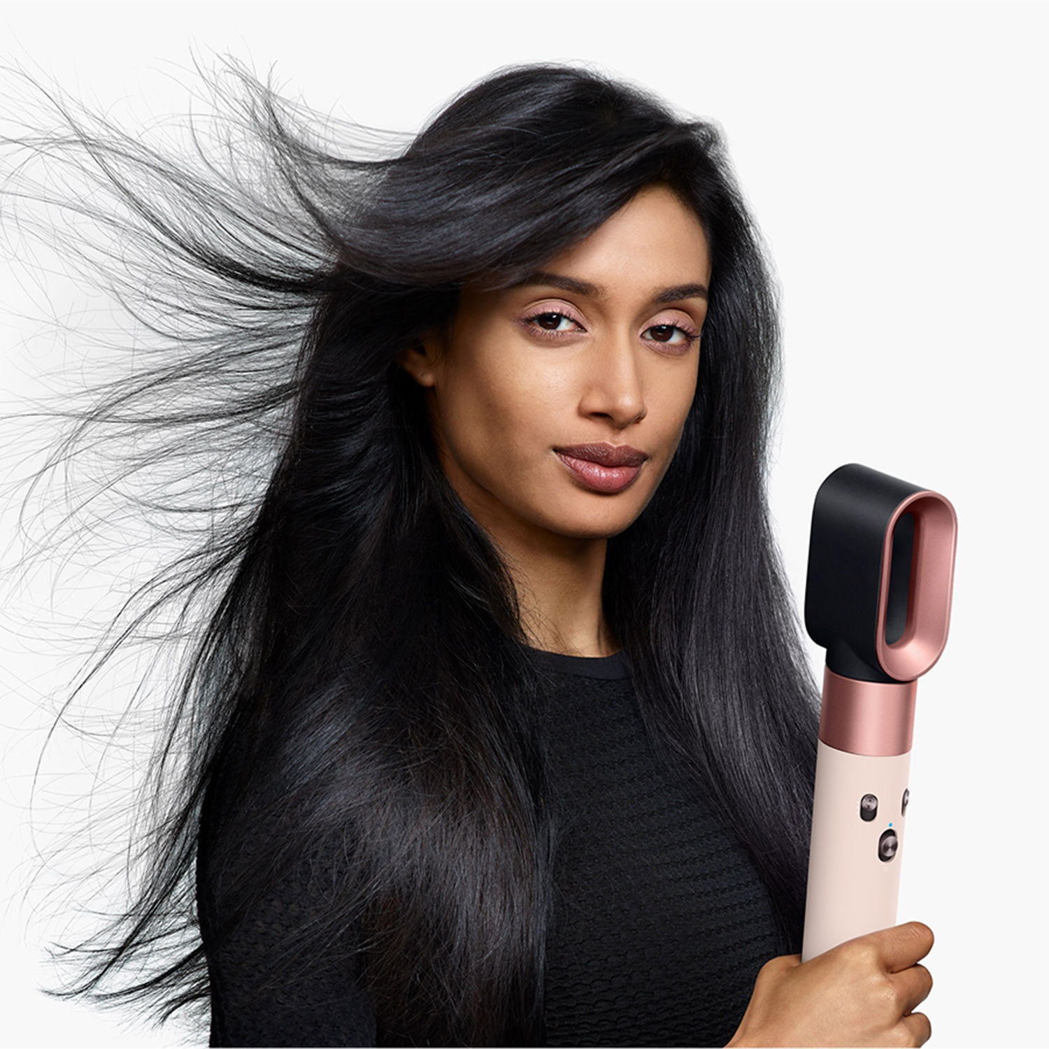 Una donna con i capelli neri tiene in mano un asciugacapelli rosa e nero. I suoi capelli sono acconciati e mossi dal vento. Indossa una camicia nera.