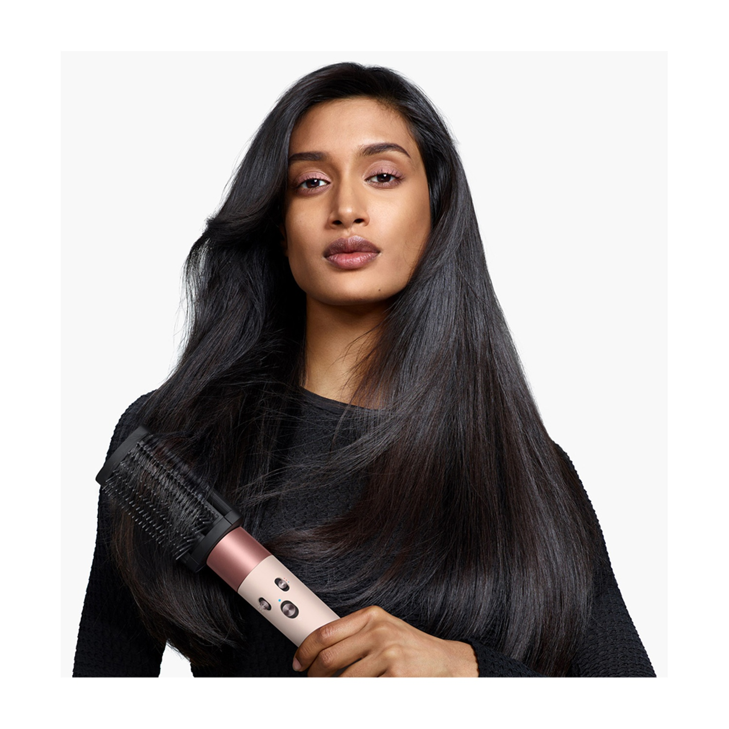 Una donna con lunghi capelli neri tiene in mano uno strumento per lo styling dei capelli. Lo strumento è nero e oro rosa. Indossa un maglione nero.
