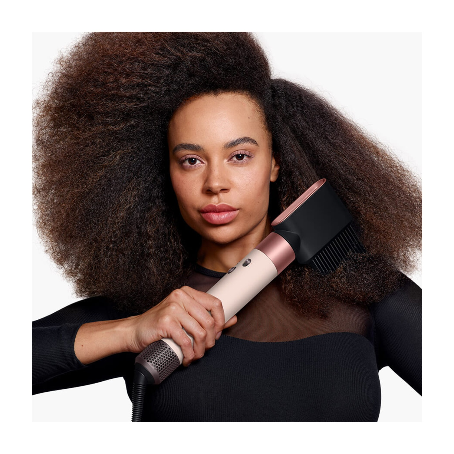 Una donna con grandi afro tiene un asciugacapelli. Indossa un top nero. L'asciugacapelli è bianco e oro rosa.