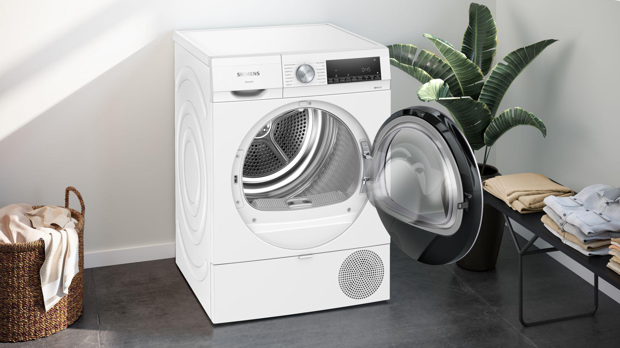 Ein weißer Siemens-Trockner mit offener Tür. Im Hintergrund eine Pflanze und gefaltete Kleidung auf einem Tisch.
