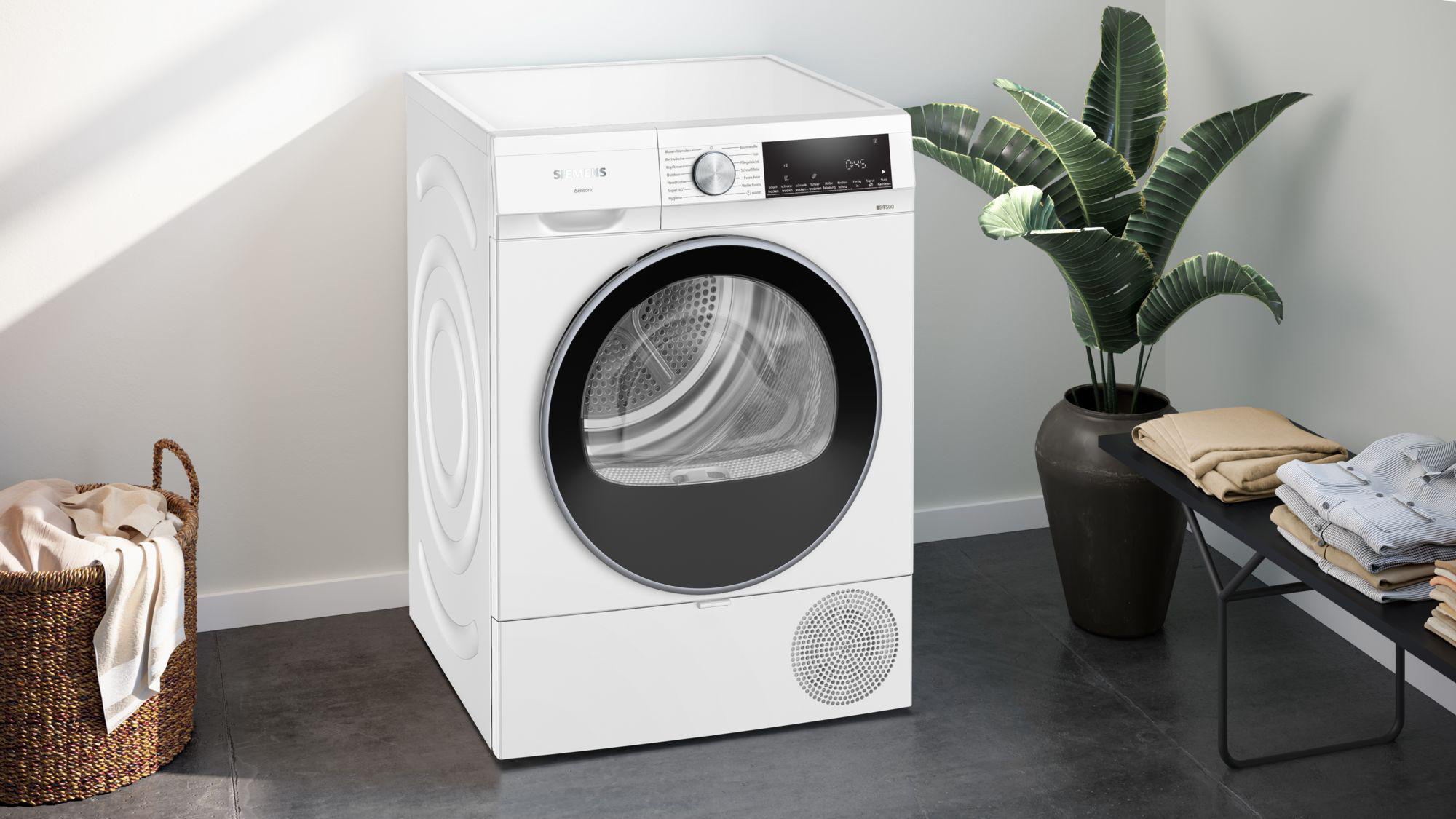 Ein weißer Siemens-Trockner mit schwarzer Tür, eine Pflanze und ein Korb mit Kleidung. Eine Bank hält gefaltete Kleidung.
