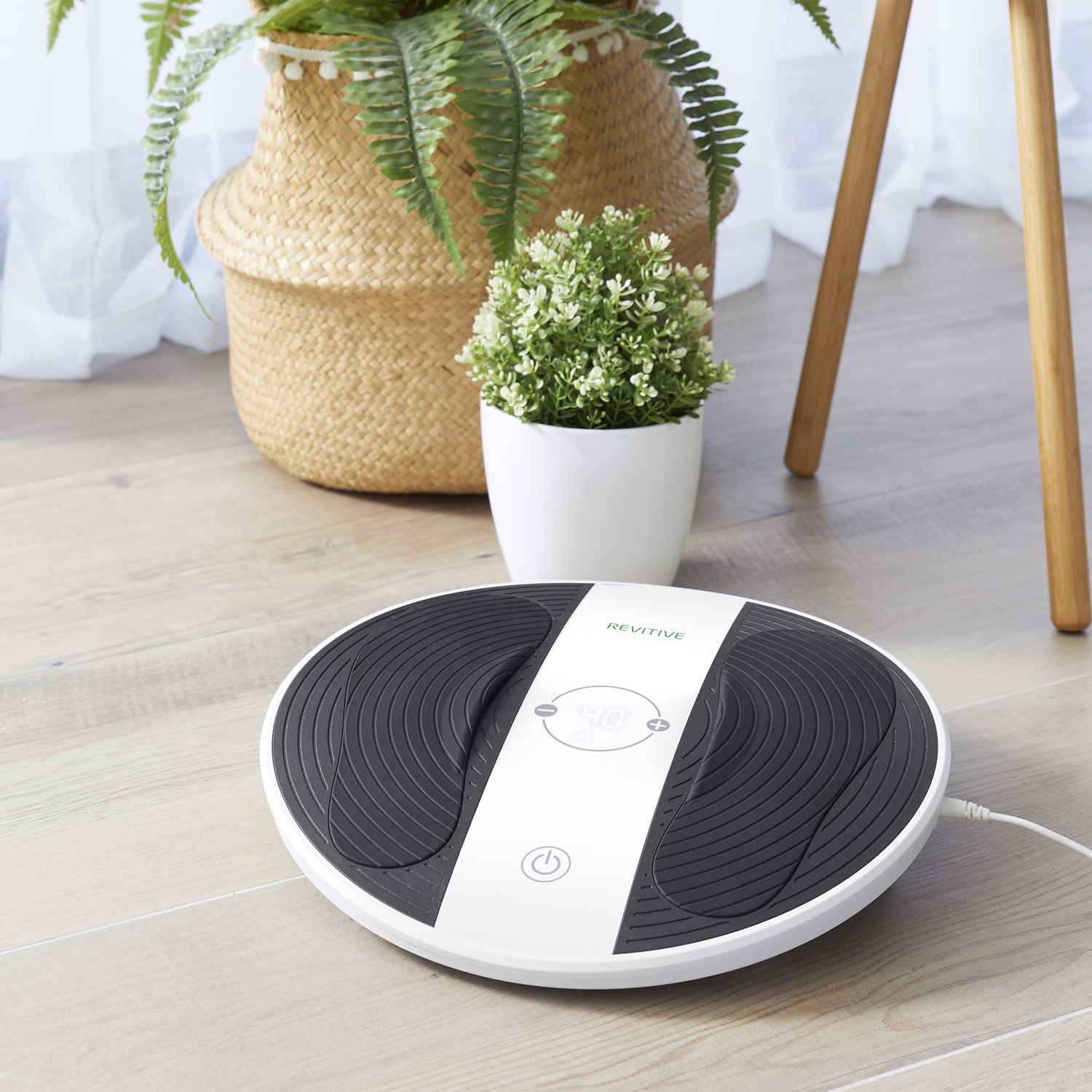 A white and black Revitive machine sits on a wooden floor. There are plants in woven baskets and pots in the background.