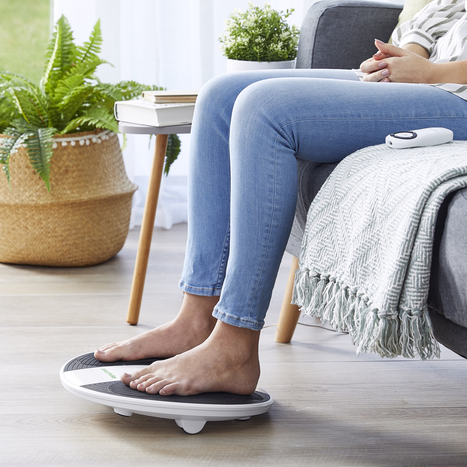 Bare feet on a balance board. A woman in jeans sits on a couch. A plant is in the background.