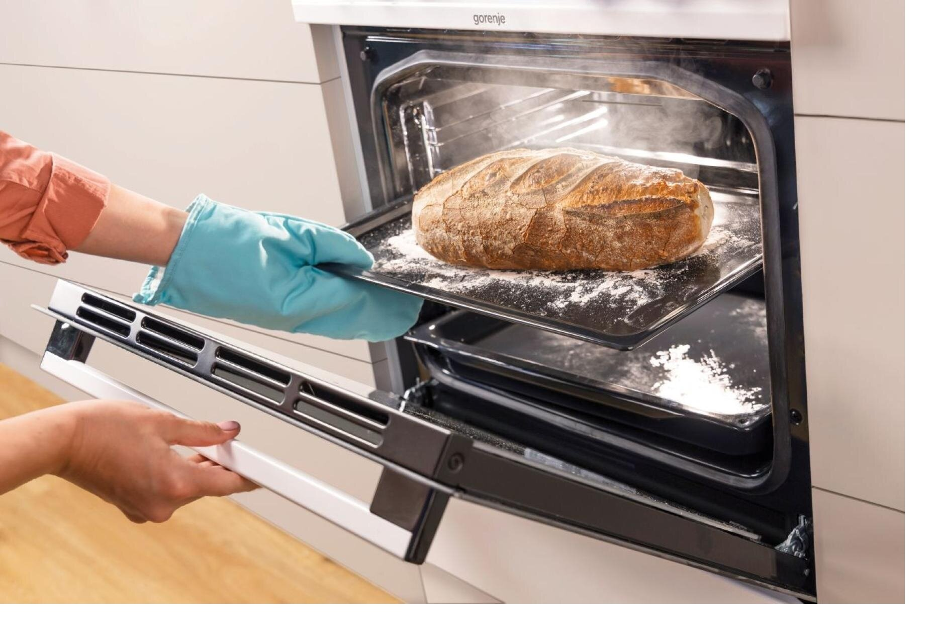 Osoba wyjmuje bochenek chleba z piekarnika za pomocą niebieskiej rękawicy kuchennej. Piekarnik jest otwarty, a chleb leży na tacy.