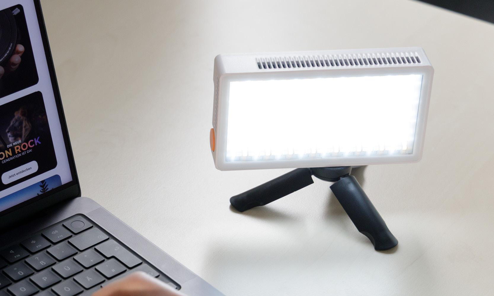 Ein helles LED-Licht auf einem kleinen Stativ steht auf einem Tisch mit einem Laptop. Das Licht ist an und beleuchtet den Bereich. Der Hintergrund ist unscharf.
