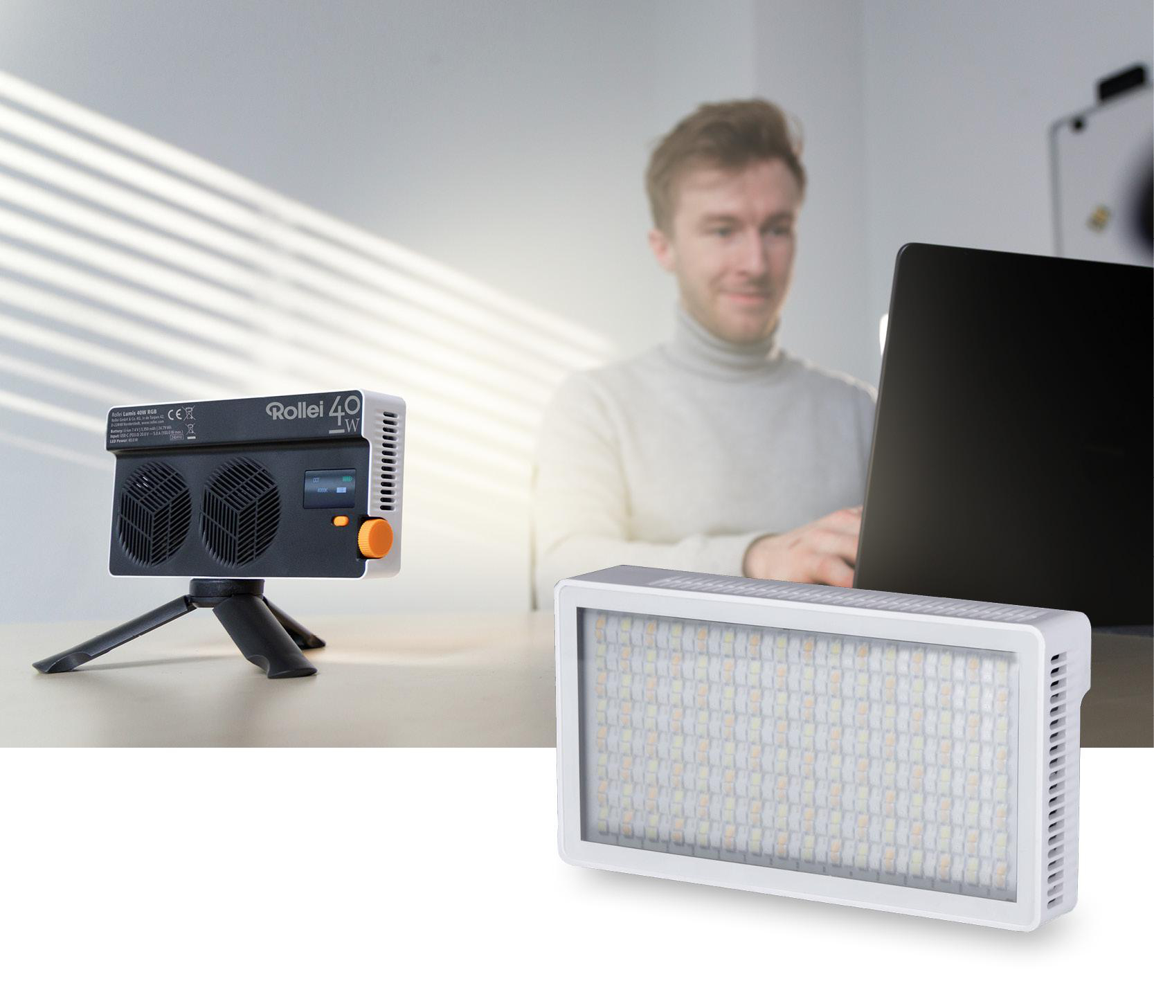 Ein Mann benutzt einen Laptop in der Nähe eines Lichts. Ein Studiolicht und ein tragbares Licht stehen auf dem Tisch. Sonnenlicht scheint aus dem Fenster.