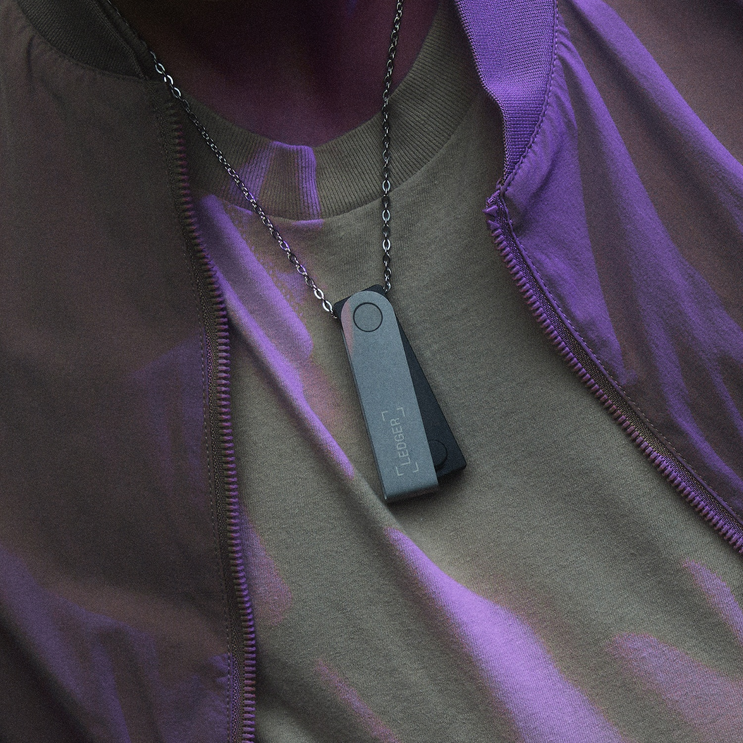Una persona indossa una collana con un dispositivo Ledger. La persona indossa una camicia verde e una giacca viola. Lo sfondo è scuro.