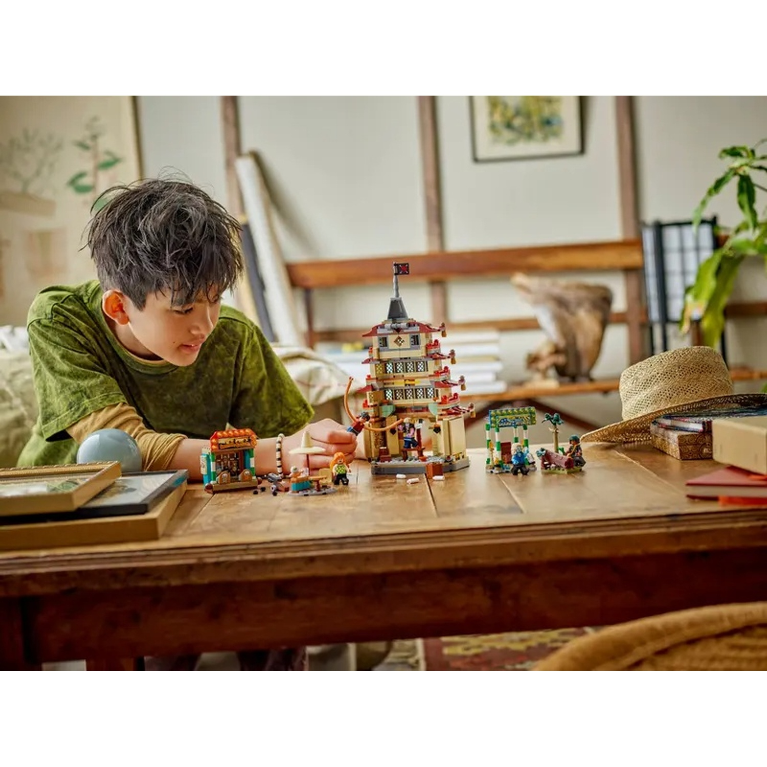 Un ragazzo gioca con costruzioni Lego su un tavolo di legno. Un cappello di paglia, libri e una pianta sullo sfondo. Il ragazzo indossa una camicia verde.