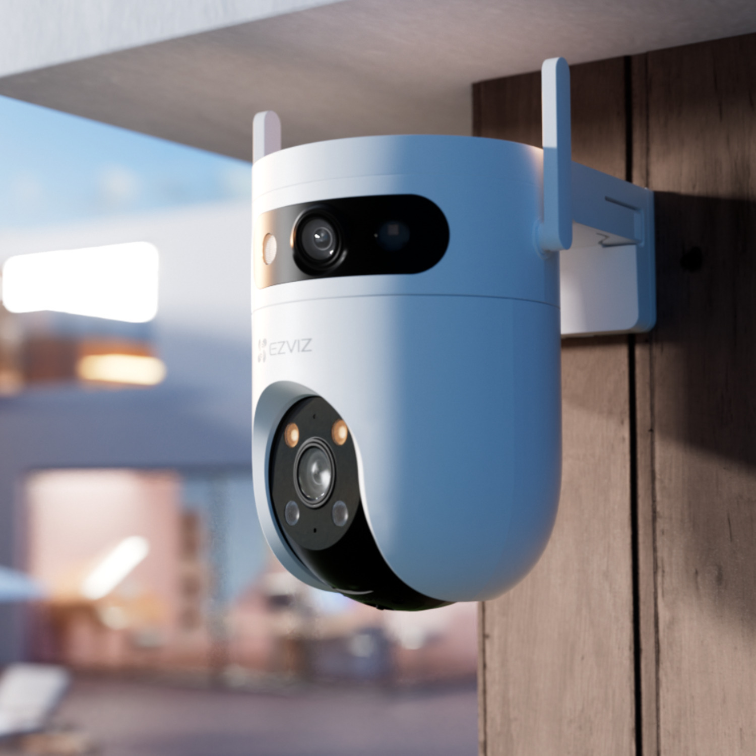 A white security camera with two antennas is mounted on a wooden surface. The background shows a blurred outdoor setting.