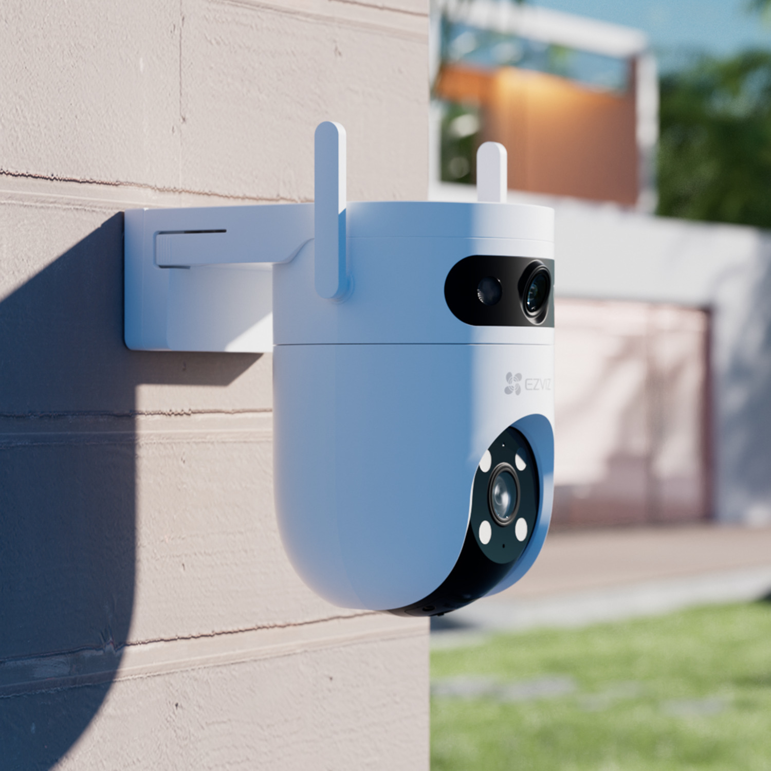 A white security camera is mounted on a light-brown wall. It has two antennas and a black lens. In the background, a house and lawn.