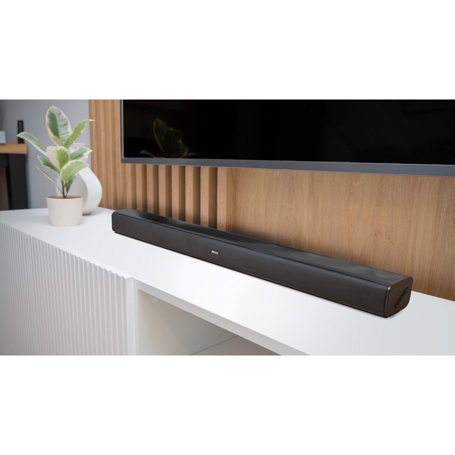 Una soundbar nera è su una mensola bianca sotto una TV. A sinistra una pianta in vaso bianco.