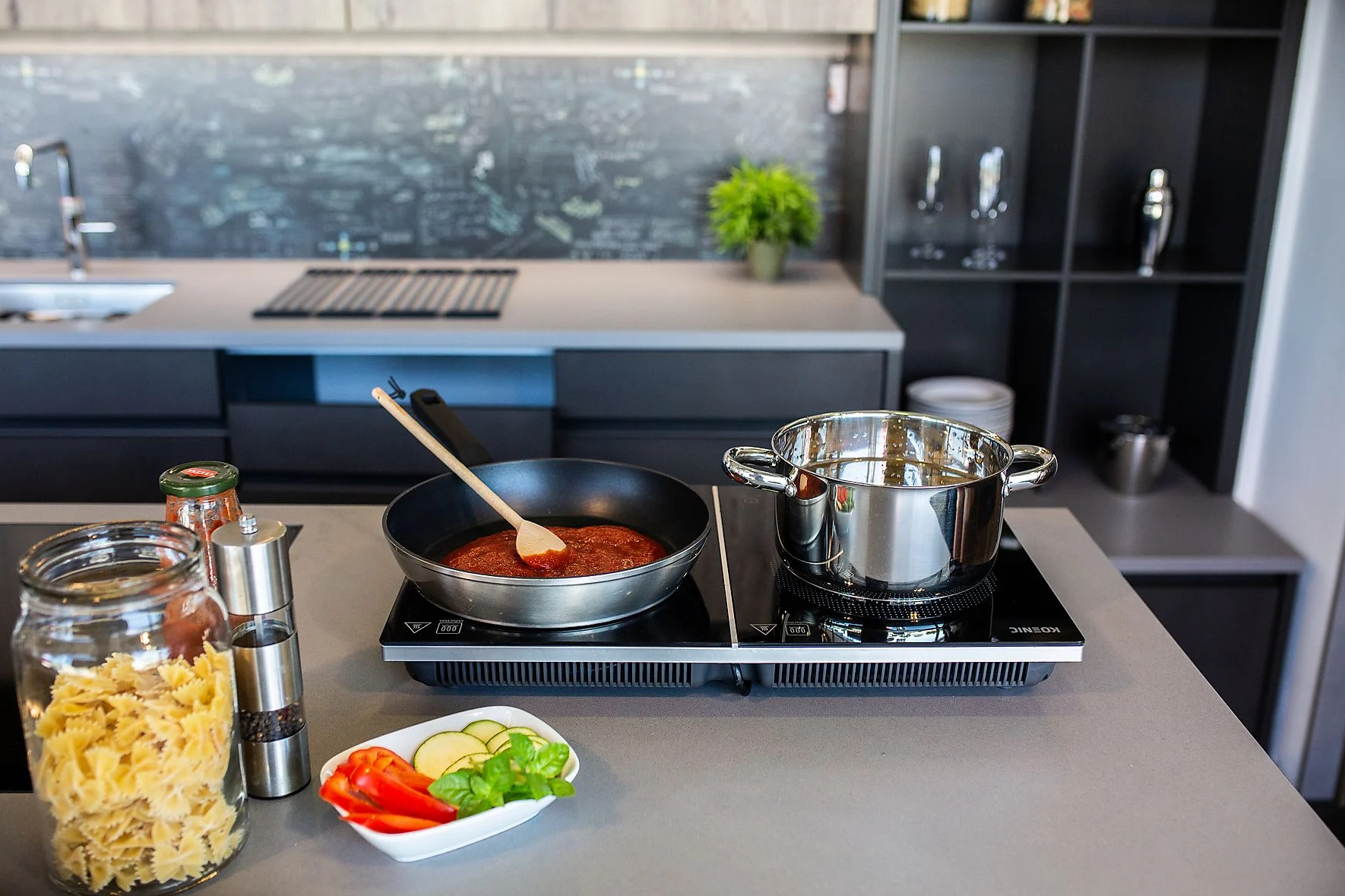 Scena kuchenna. Patelnia z sosem, garnek, słoik makaronu, warzywa i przyprawy na blacie. Czarna płyta grzewcza i tło.