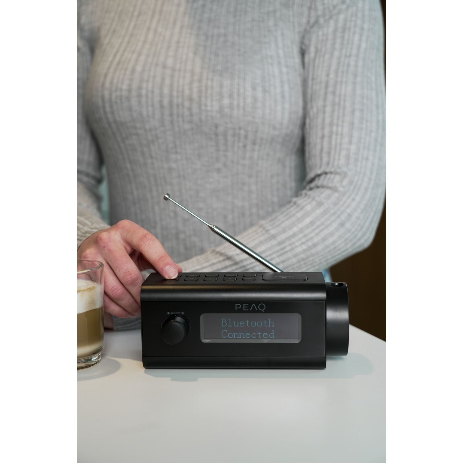 Mani su una radio nera con scritto 'Bluetooth Connected'. Un bicchiere di caffè sul tavolo. La persona indossa un maglione grigio.