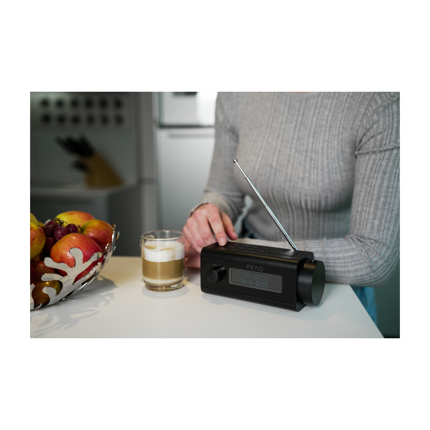 Una persona tocca una radio nera su un tavolo bianco. Un bicchiere di latte macchiato e una fruttiera sono anche sul tavolo.