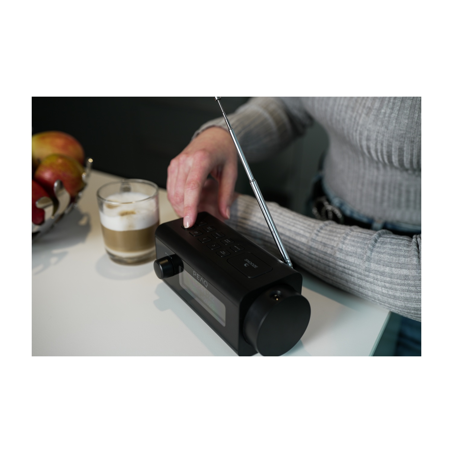 Una mano preme un pulsante su una radio nera. Accanto, un bicchiere di latte macchiato e una ciotola di mele su un tavolo bianco.