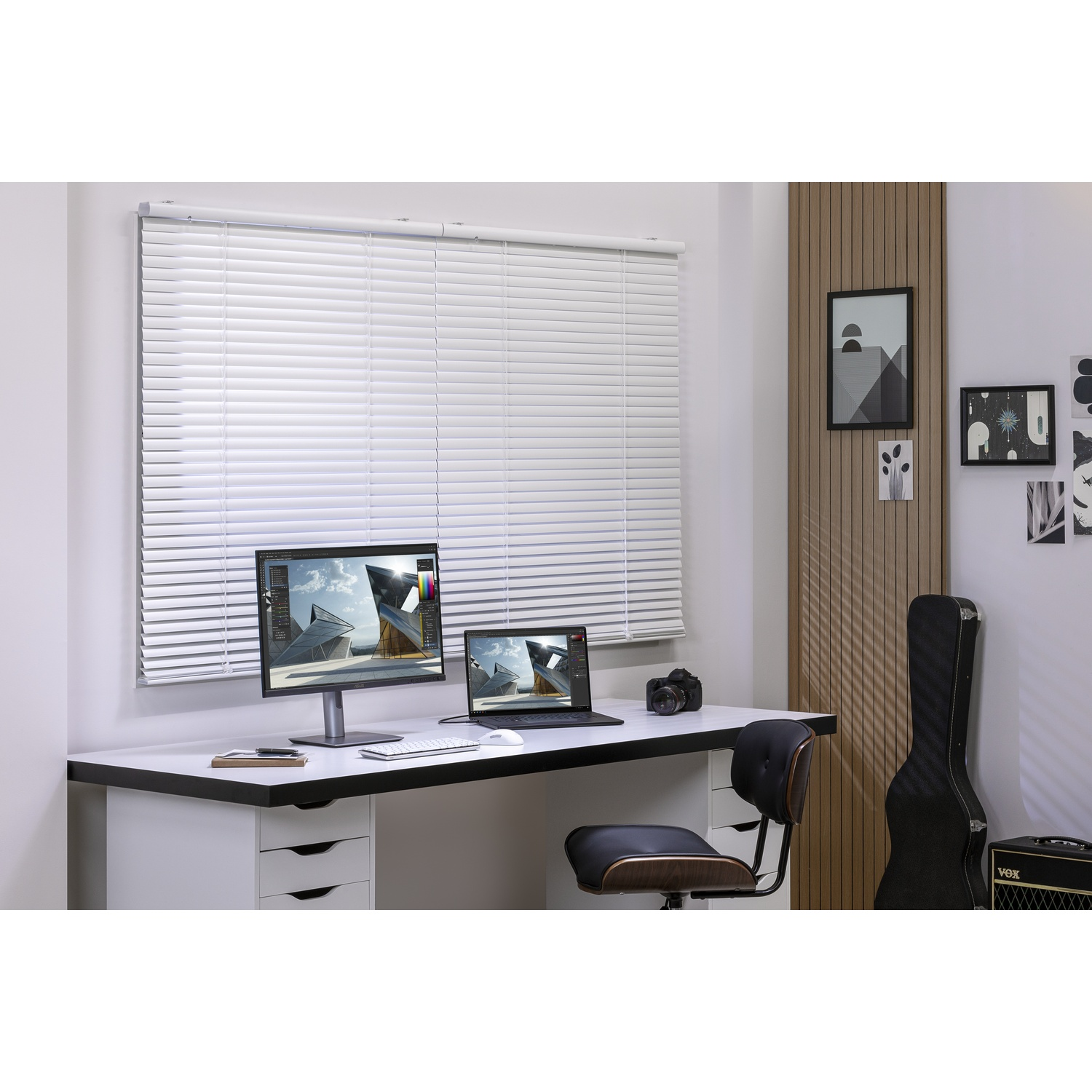 Un espace de travail moderne avec un ordinateur, un ordinateur portable, un appareil photo et un étui de guitare. La pièce a une fenêtre avec des stores et de l'art mural décoratif.