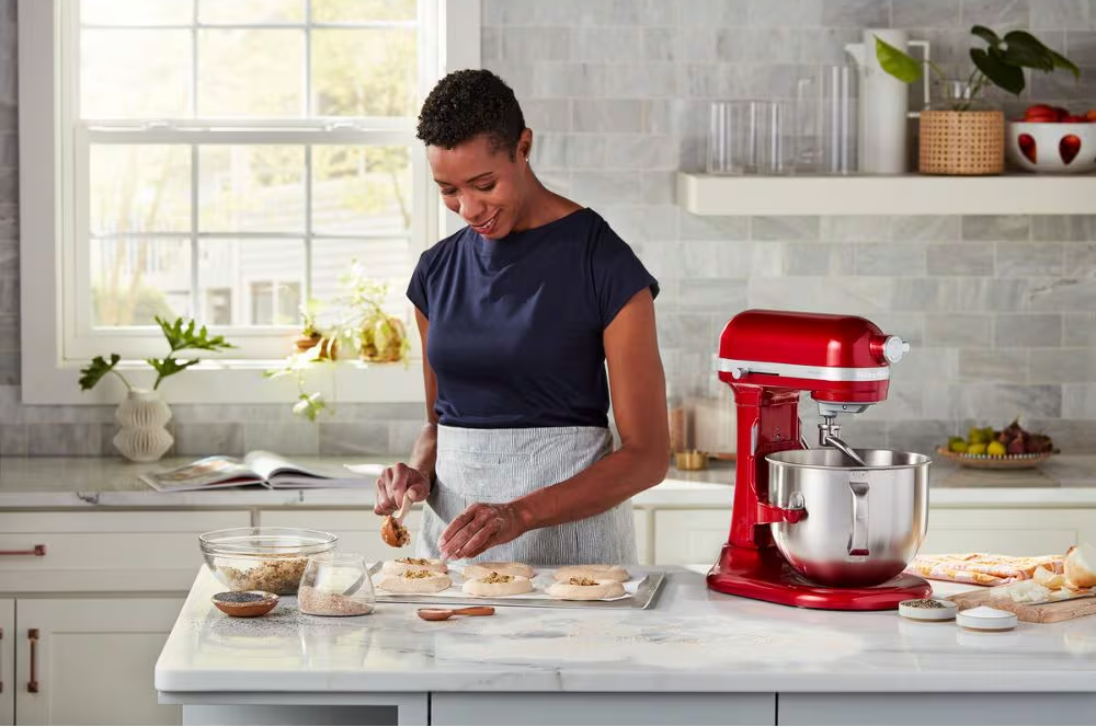 Una donna in cucina prepara del cibo. Un mixer rosso è sulla destra. Gli ingredienti sono sul bancone e la luce del sole entra da una finestra.