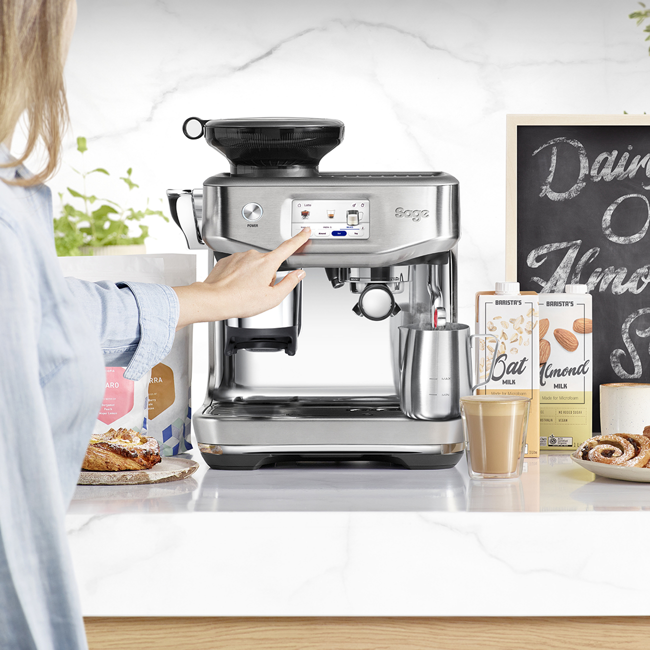 Eine Frau bedient eine Espressomaschine. Auf der Theke stehen Mandelmilch, Gebäck und Kaffee. Ein Tafelschild ist vorhanden.