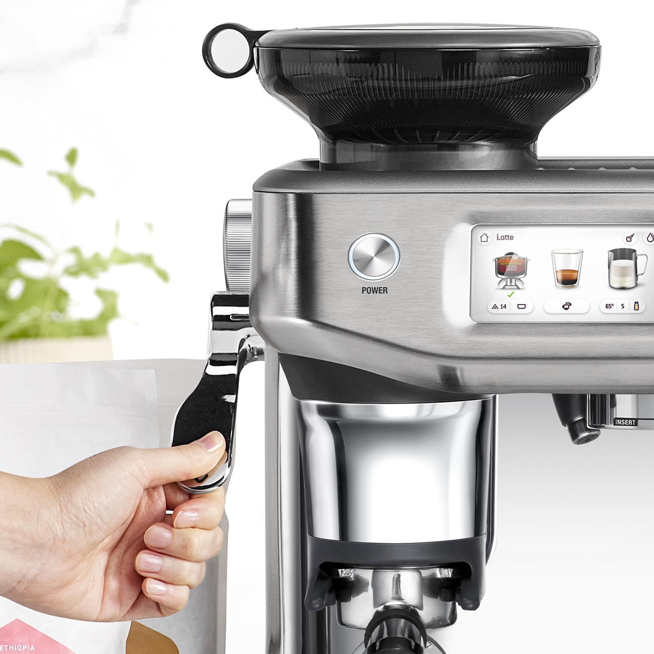 Eine silberne Espressomaschine mit einem schwarzen Bohnenbehälter. Eine Hand hält einen Hebel. Ein Netzschalter ist sichtbar.