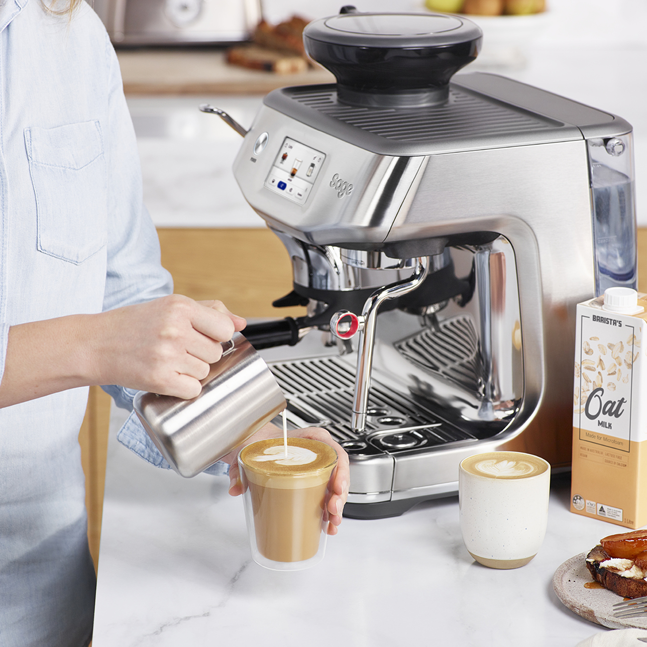 Una persona versa il latte nel caffè. Una macchina da caffè sullo sfondo, con latte d'avena e un muffin sul tavolo.