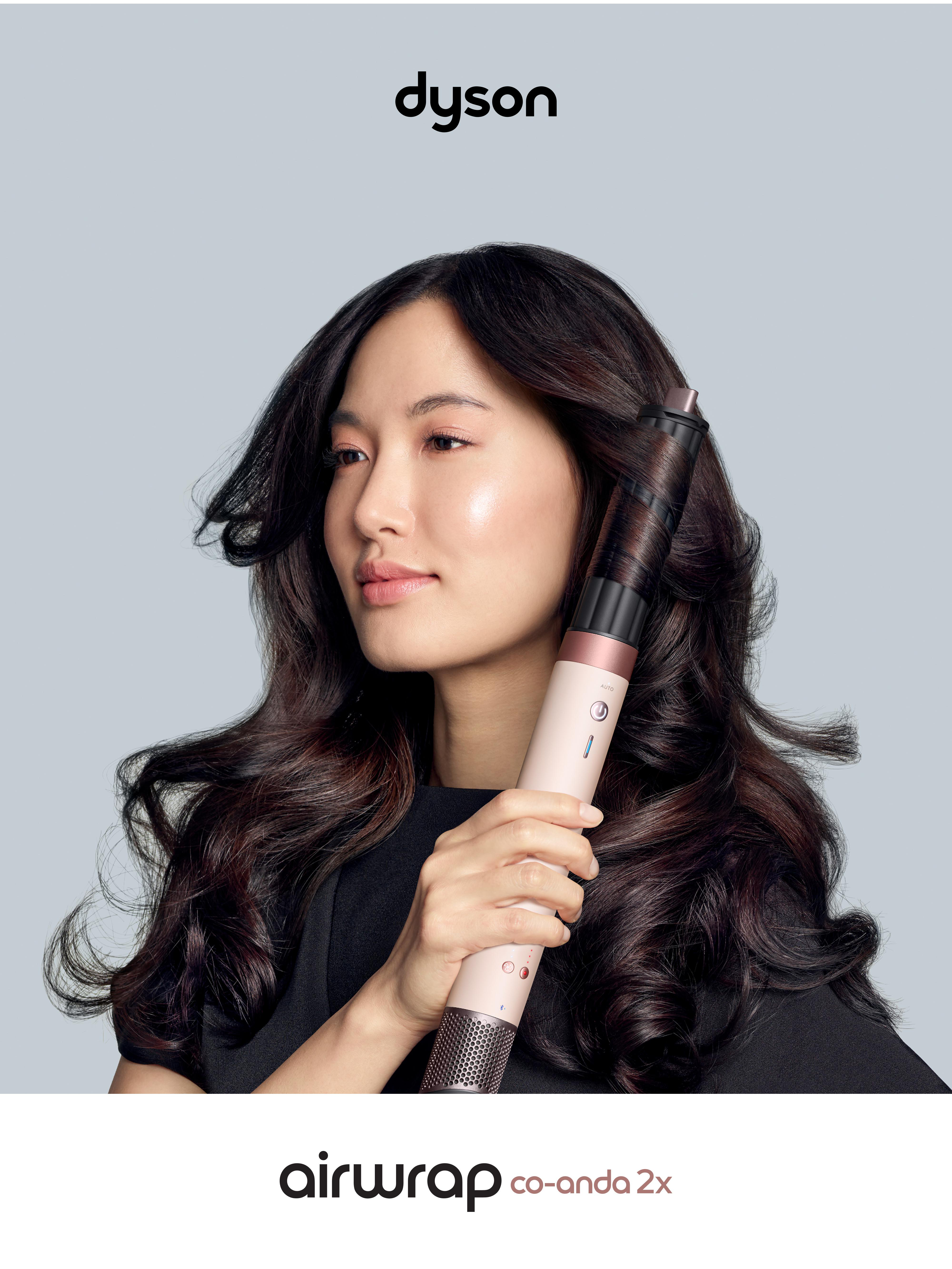 Frau benutzt Dyson Airwrap. Sie hält den Styler an ihr dunkles Haar, das in Wellen gestylt ist. Der Hintergrund ist grau.