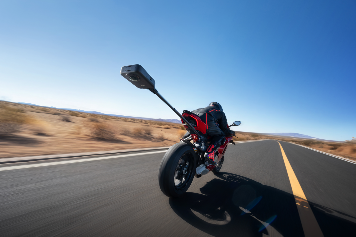 Un motociclista monta una moto roja y negra en una carretera. Hay una cámara montada en la moto. El fondo muestra desierto y cielo azul.
