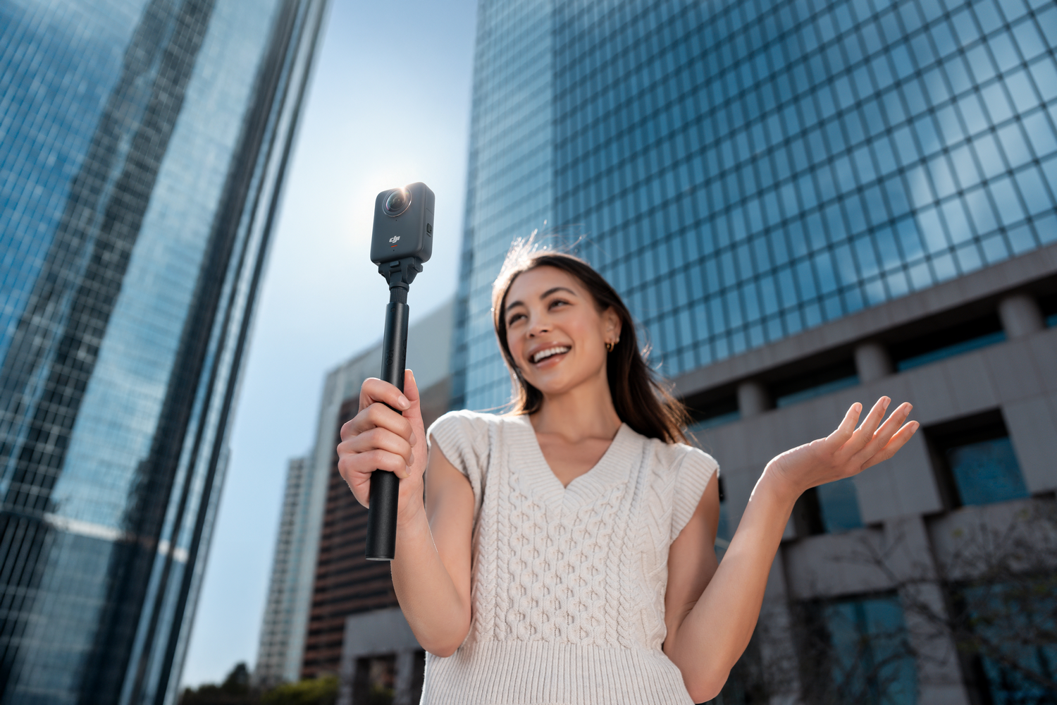 Mujer sosteniendo una cámara en un palo selfie, sonriendo. Edificios altos al fondo, día soleado.