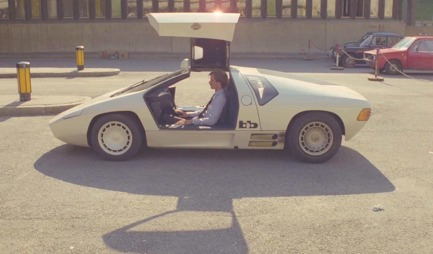 Ein Mann sitzt in einem cremefarbenen futuristischen Auto mit offener Flügeltür. Im Hintergrund parken weitere Autos.