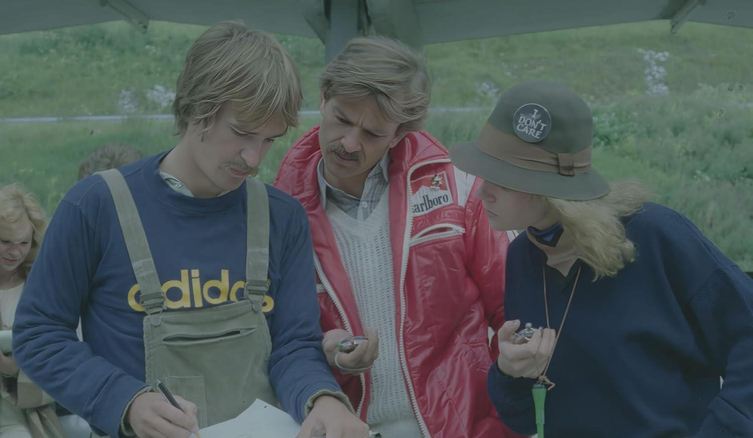 Drei Personen betrachten ein Papier. Einer trägt einen Hut mit der Aufschrift "I DON'T CARE". Ein anderer trägt eine rote Jacke mit "Marlboro".