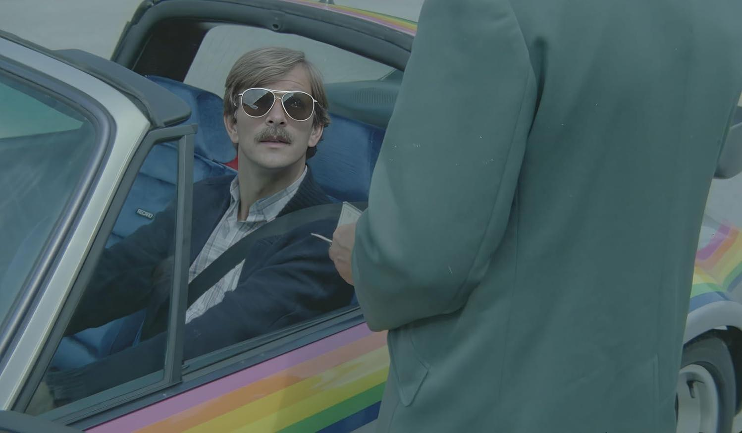 Ein Mann mit Sonnenbrille sitzt in einem Auto. Ein anderer Mann in einem Anzug steht draußen. Das Auto ist in Regenbogenfarben lackiert.